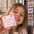 Smiling girl holding a Minigallery magnet framing her 'I love you mom' drawing, with more framed drawings on the fridge behind her